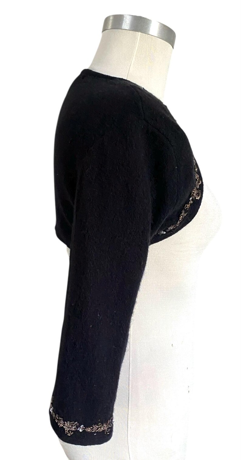 May include: Black bolero sweater with three-quarter sleeves. The sweater has a rounded neckline and is adorned with a decorative, beaded trim along the sleeve cuffs and the front opening. The garment is displayed on a white mannequin.