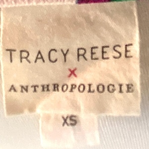 May include: A close-up of a tag with the text "TRACY REESE x ANTHROPOLOGIE" in black letters. Below the text is the size "XS". The tag is a light beige color and is attached to a white fabric.