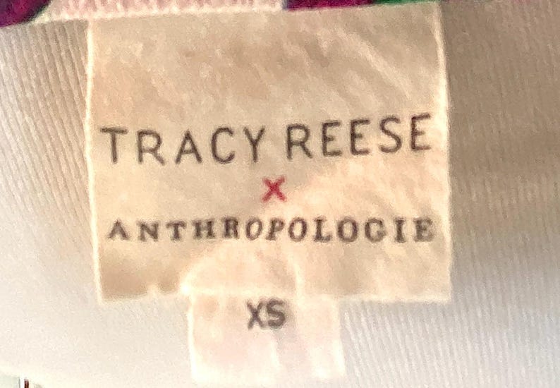 May include: A close-up of a tag with the text "TRACY REESE x ANTHROPOLOGIE" in black letters. Below the text is the size "XS". The tag is a light beige color and is attached to a white fabric.