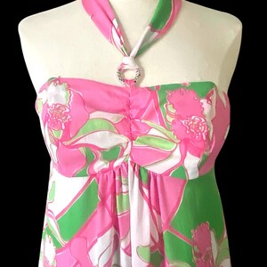 Op de afbeelding: Een strapless jurk met halternek, met een levendig bloemenpatroon in roze, groen en wit. De halterband is versierd met een sprankelende, cirkelvormige versiering. De jurk wordt op een mannequin getoond.