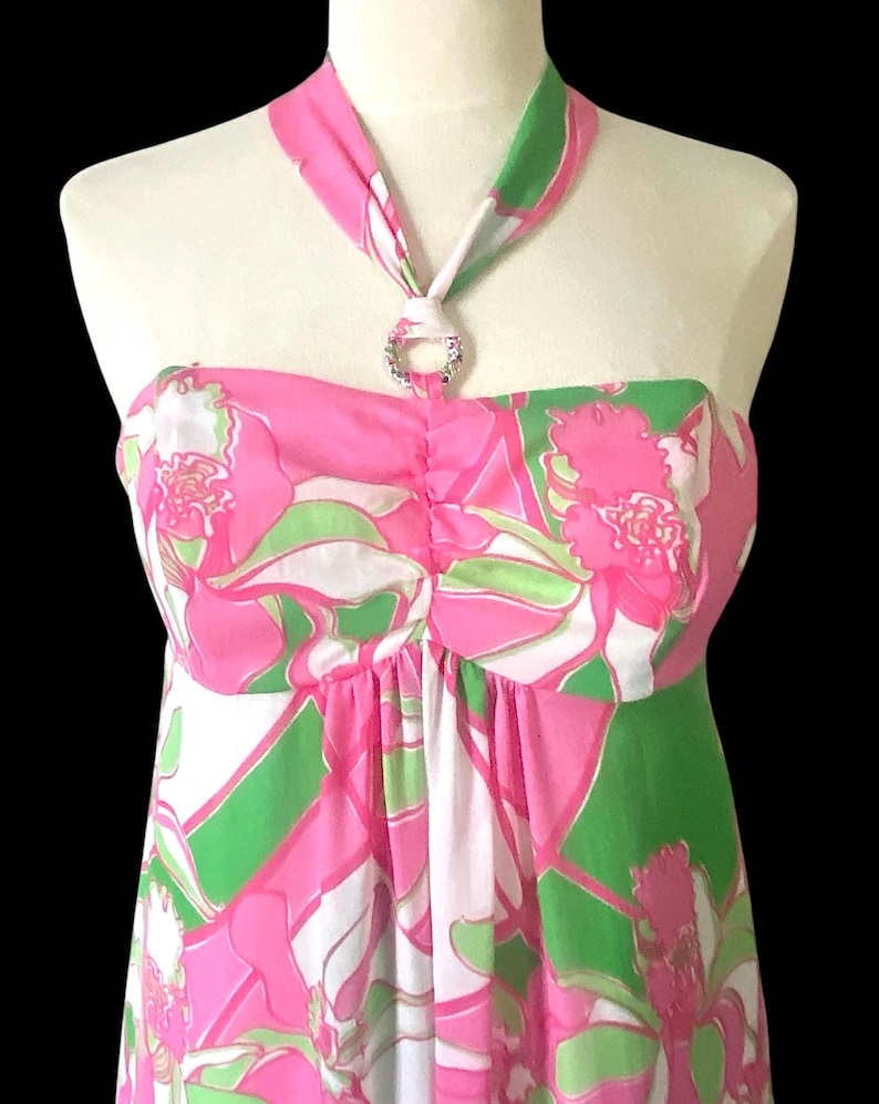 Op de afbeelding: Een strapless jurk met halternek, met een levendig bloemenpatroon in roze, groen en wit. De halterband is versierd met een sprankelende, cirkelvormige versiering. De jurk wordt op een mannequin getoond.
