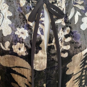 Puede incluir: Un vestido de terciopelo negro con estampado floral en tonos marrones, beige y morado. El vestido tiene un escote de ojo de cerradura con cierre de lazo negro.