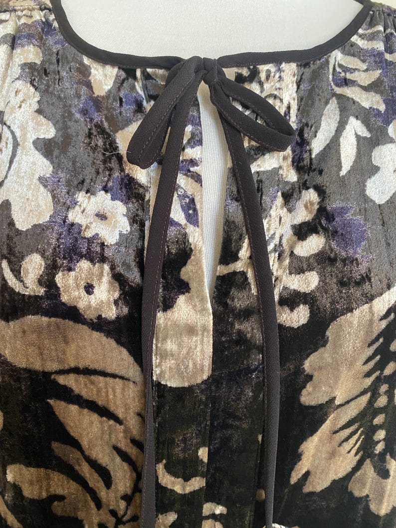 Puede incluir: Un vestido de terciopelo negro con estampado floral en tonos marrones, beige y morado. El vestido tiene un escote de ojo de cerradura con cierre de lazo negro.