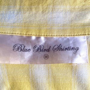 K&ouml;nnte beinhalten: Ein wei&szlig;es Etikett mit dem Text "Blue Bird Shirting" in schwarzer Kursivschrift und einer Gr&ouml;&szlig;e "M" in einem Kreis. Das Etikett befindet sich auf einem gelb-wei&szlig; gestreiften Stoff.