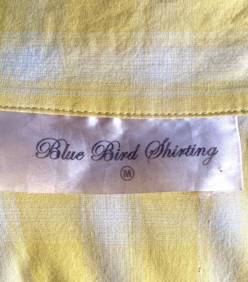 K&ouml;nnte beinhalten: Ein wei&szlig;es Etikett mit dem Text "Blue Bird Shirting" in schwarzer Kursivschrift und einer Gr&ouml;&szlig;e "M" in einem Kreis. Das Etikett befindet sich auf einem gelb-wei&szlig; gestreiften Stoff.