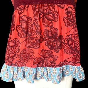 May include: A sleeveless top with a red background and large, dark brown floral patterns. The top has a burgundy lace neckline and a blue and white patterned ruffle at the hem. The overall style is bohemian.