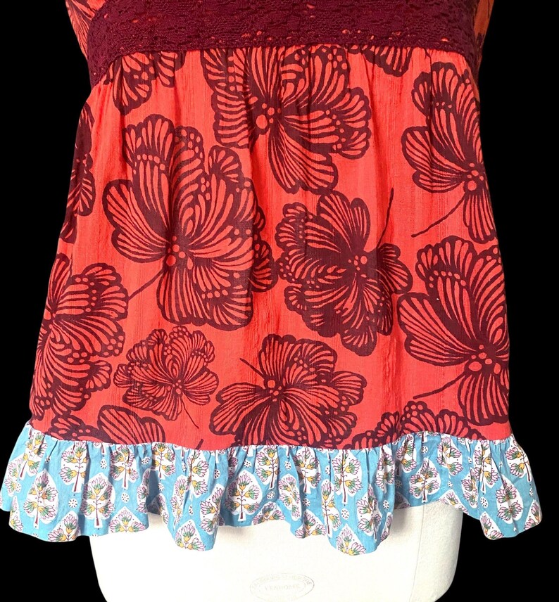 May include: A sleeveless top with a red background and large, dark brown floral patterns. The top has a burgundy lace neckline and a blue and white patterned ruffle at the hem. The overall style is bohemian.