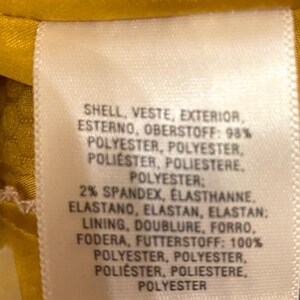 Op de afbeelding: Een close-up van een wit kledinglabel met tekst in meerdere talen. Het label geeft de samenstelling aan: 98% polyester voor de buitenkant, 2% spandex en een voering van 100% polyester. De achtergrond is gele stof.