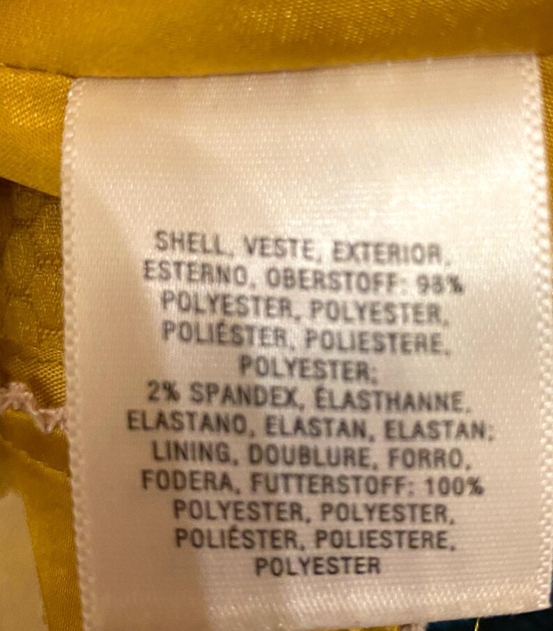 Op de afbeelding: Een close-up van een wit kledinglabel met tekst in meerdere talen. Het label geeft de samenstelling aan: 98% polyester voor de buitenkant, 2% spandex en een voering van 100% polyester. De achtergrond is gele stof.
