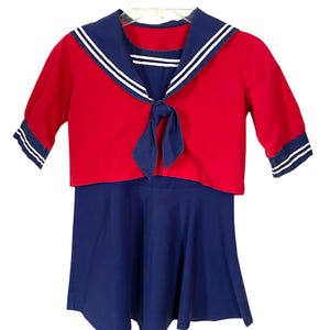 May include: A vintage sailor-style outfit featuring a red top with a navy blue collar and sleeves, accented with white stripes. The top has a tie detail at the neckline and is paired with a navy blue skirt. The outfit is displayed on a white hanger.