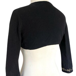 May include: Black bolero sweater with three-quarter sleeves. The sweater has a rounded neckline and is adorned with an ornate, silver-toned floral design at the cuffs. The garment is displayed on a white mannequin.