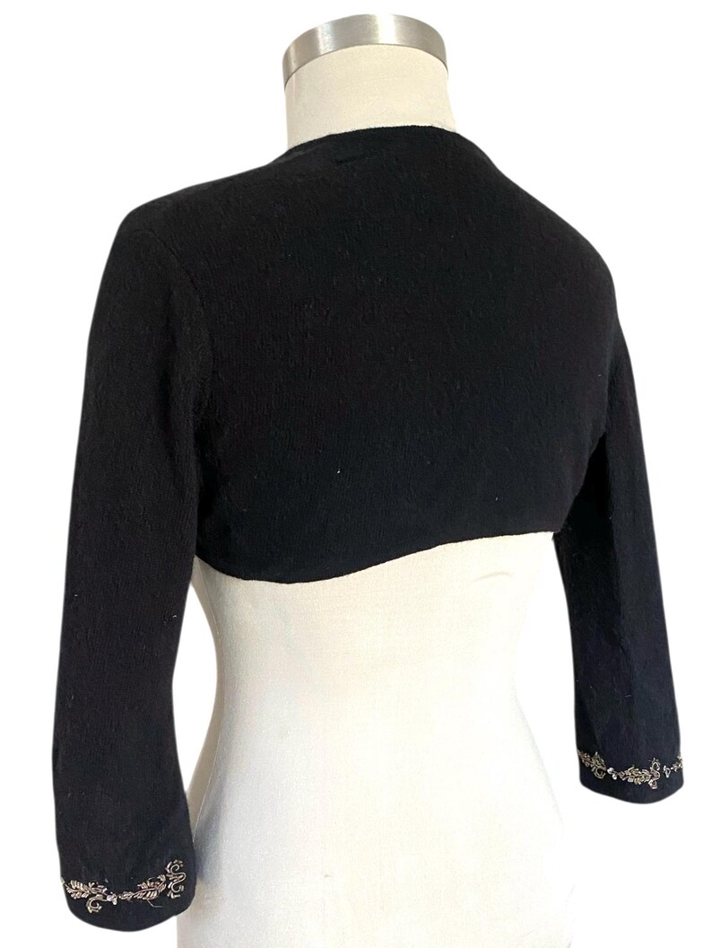 May include: Black bolero sweater with three-quarter sleeves. The sweater has a rounded neckline and is adorned with an ornate, silver-toned floral design at the cuffs. The garment is displayed on a white mannequin.