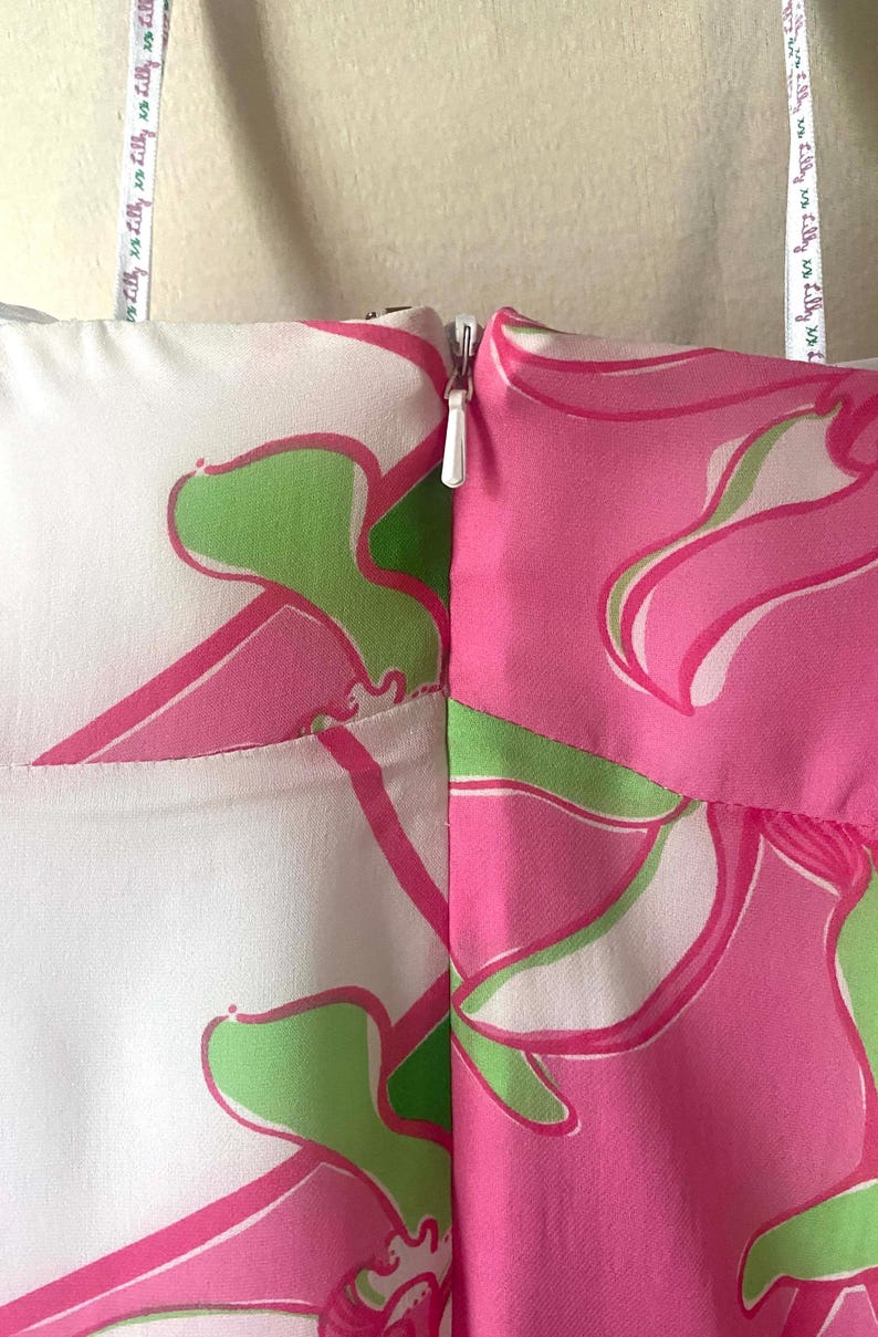 Op de afbeelding: Close-up van een kledingstuk met een witte rits. De stof heeft een bloemenpatroon in roze en groen op een witte achtergrond. Een meetlint met tekst is zichtbaar.