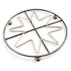May include: Circular metal trivet with a starburst design. The trivet features a black outer ring and intersecting lines, with a gold-colored star in the center. Small gold-colored ball feet elevate the trivet.