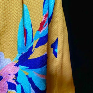 Op de afbeelding: Close-up van een gele stof met een textuurpatroon. De stof heeft een groot bloemmotief in blauwe, roze en rode tinten. De mouw van een kledingstuk is zichtbaar aan de rechterkant, in een effen gele kleur.