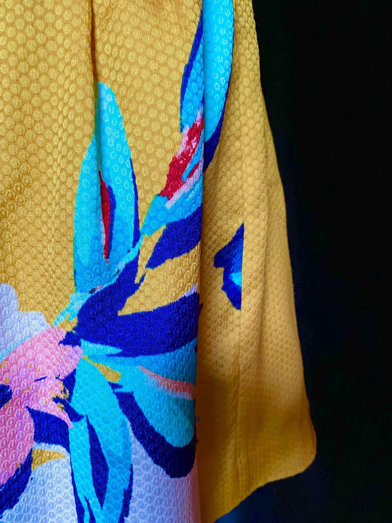 Op de afbeelding: Close-up van een gele stof met een textuurpatroon. De stof heeft een groot bloemmotief in blauwe, roze en rode tinten. De mouw van een kledingstuk is zichtbaar aan de rechterkant, in een effen gele kleur.