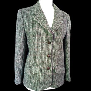 May include: A blazer featuring a green, gray, and brown herringbone pattern with a notched lapel, three dark buttons down the front, and two flap pockets. The garment is constructed with long sleeves and a structured fit, showcasing a textured weave.