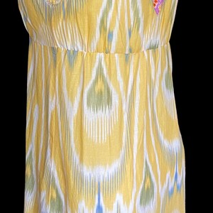 May include: Yellow sleeveless dress with a V-neckline and a floral patterned trim. The dress features a white, blue, and green ikat pattern. The dress is displayed on a mannequin against a black background.