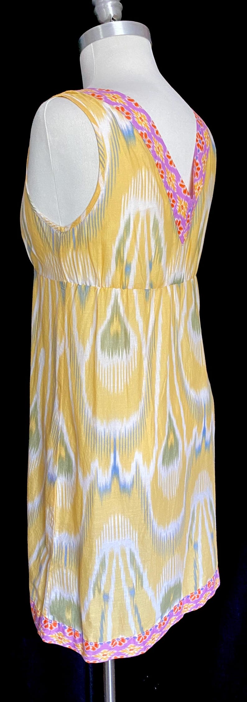 May include: Yellow sleeveless dress with a V-neckline and a floral patterned trim. The dress features a white, blue, and green ikat pattern. The dress is displayed on a mannequin against a black background.