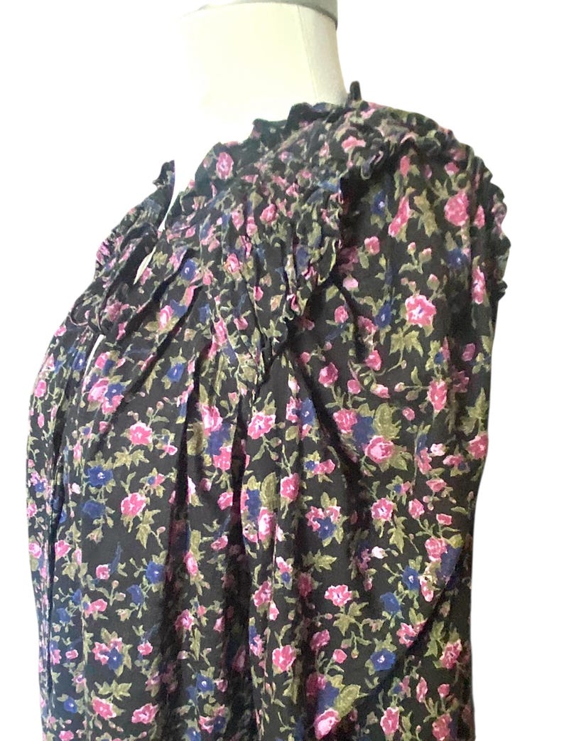 May include: A black floral print blouse with pink, blue, and green flowers. The blouse has a ruffled neckline and sleeves, and a tie detail at the neck. The fabric appears to be lightweight and flowy.