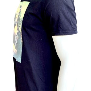 May include: Black short-sleeved t-shirt with a rectangular graphic on the front. The graphic appears to be a painting with yellow and green tones. The shirt is displayed on a mannequin.