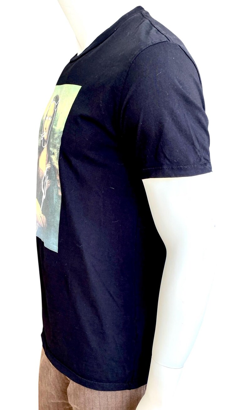 May include: Black short-sleeved t-shirt with a rectangular graphic on the front. The graphic appears to be a painting with yellow and green tones. The shirt is displayed on a mannequin.