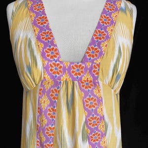 May include: A sleeveless yellow dress with a V-neckline. The dress features a colorful ikat pattern with vertical stripes in yellow, white, and green. The neckline and straps are adorned with a purple floral design. The dress is displayed on a mannequin.