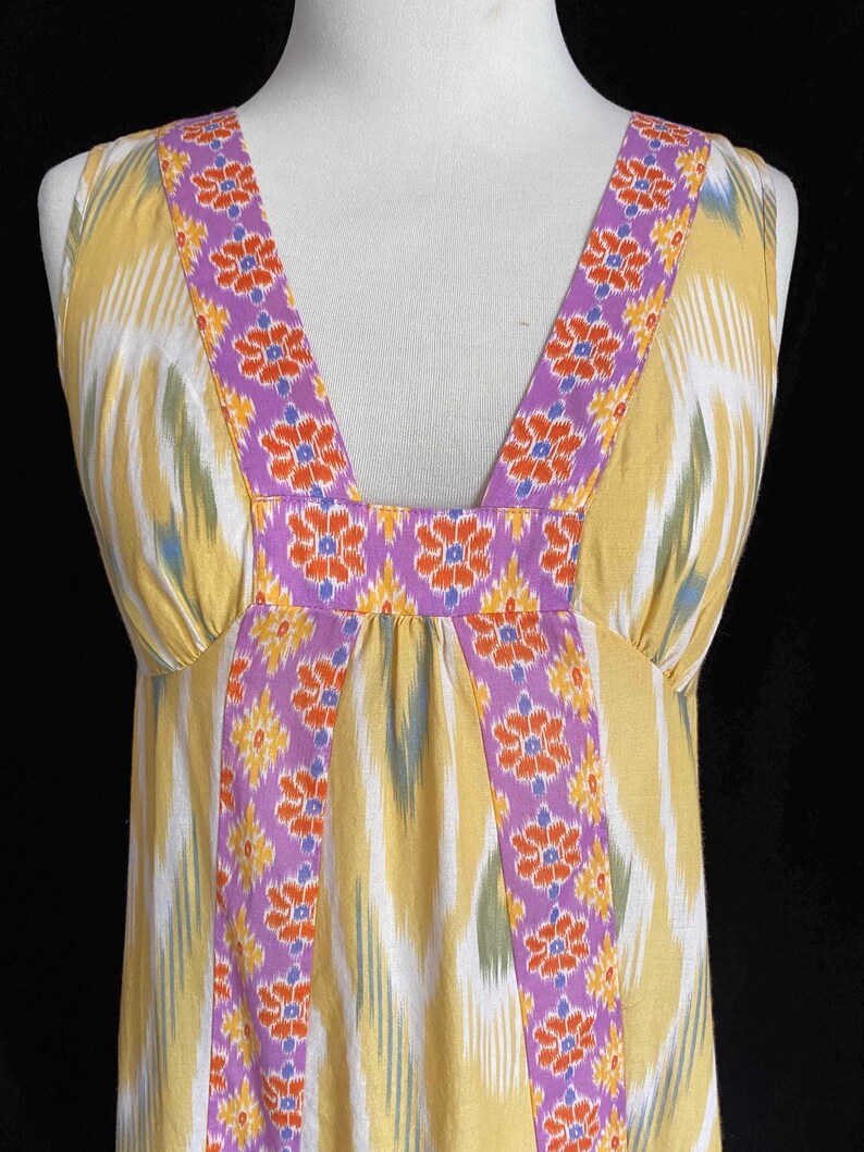 May include: A sleeveless yellow dress with a V-neckline. The dress features a colorful ikat pattern with vertical stripes in yellow, white, and green. The neckline and straps are adorned with a purple floral design. The dress is displayed on a mannequin.