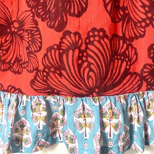 May include: Close-up of a garment featuring a vibrant coral fabric adorned with large, dark burgundy floral patterns. The bottom edge has a light blue ruffle with a repeating tree and floral design.