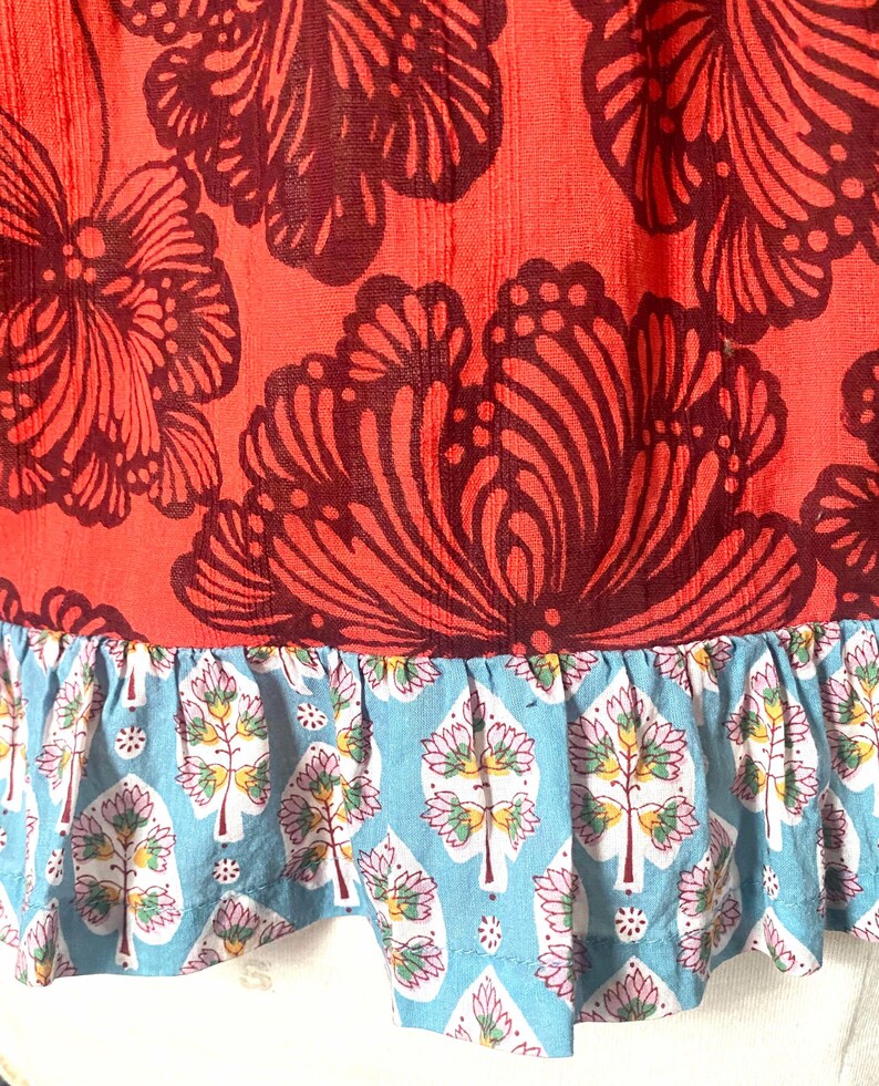 May include: Close-up of a garment featuring a vibrant coral fabric adorned with large, dark burgundy floral patterns. The bottom edge has a light blue ruffle with a repeating tree and floral design.
