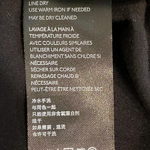 以下が含まれることがあります： 黒い布製のタグで、白い文字で衣類のお手入れ方法が記載されています。タグには、英語、フランス語、中国語での洗濯、漂白、乾燥、アイロンがけの説明が含まれています。タグは黒い衣服に付いています。