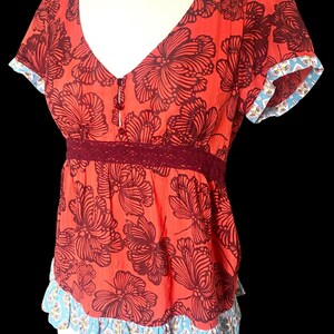 May include: A red floral print top with a V-neckline and short sleeves. The top features a dark burgundy floral pattern, a burgundy lace waist detail, and blue and white patterned trim on the sleeves and hem.