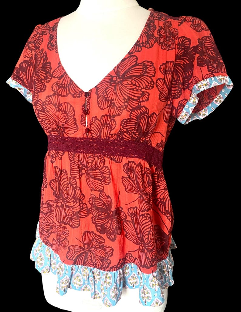 May include: A red floral print top with a V-neckline and short sleeves. The top features a dark burgundy floral pattern, a burgundy lace waist detail, and blue and white patterned trim on the sleeves and hem.