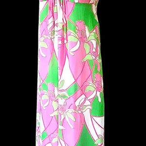 Op de afbeelding: Een lange halterjurk met een bloemenprint. De jurk heeft een roze, groene en witte print, een halterhals en een decoratieve ring. De jurk wordt gepresenteerd op een mannequin tegen een zwarte achtergrond.