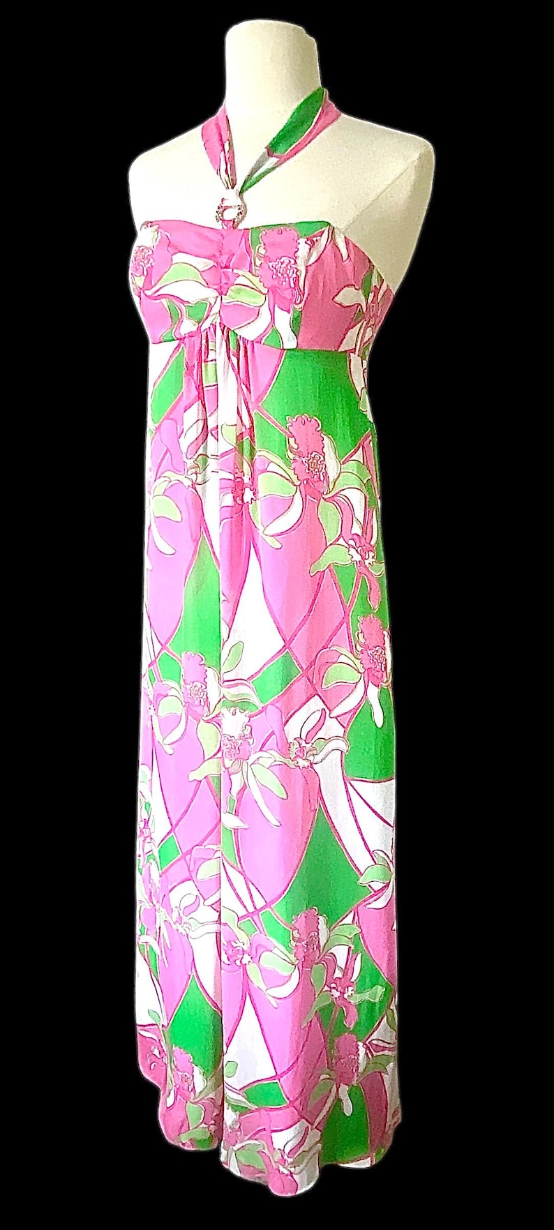 Op de afbeelding: Een lange halterjurk met een bloemenprint. De jurk heeft een roze, groene en witte print, een halterhals en een decoratieve ring. De jurk wordt gepresenteerd op een mannequin tegen een zwarte achtergrond.