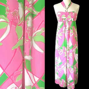 Op de afbeelding: Een maxi-jurk met halternek met een levendig bloemenpatroon in roze, groen en wit. De jurk heeft een getailleerd lijfje en een zwierige rok. De stof heeft een glanzende glans en het ontwerp doet denken aan vintage resortkleding.