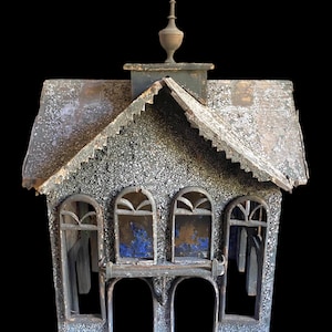 May include: A weathered wooden dollhouse with a dark gray, textured exterior finish. The structure features a gabled roof with a central finial, four arched window frames, and a small balcony. The surface shows signs of age and wear.