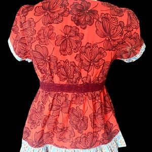May include: A red floral top with a dark brown floral pattern. The top features short sleeves with a blue and white patterned trim, a burgundy lace waist detail, and a ruffled hem with a blue and white pattern. The top is displayed on a white mannequin.