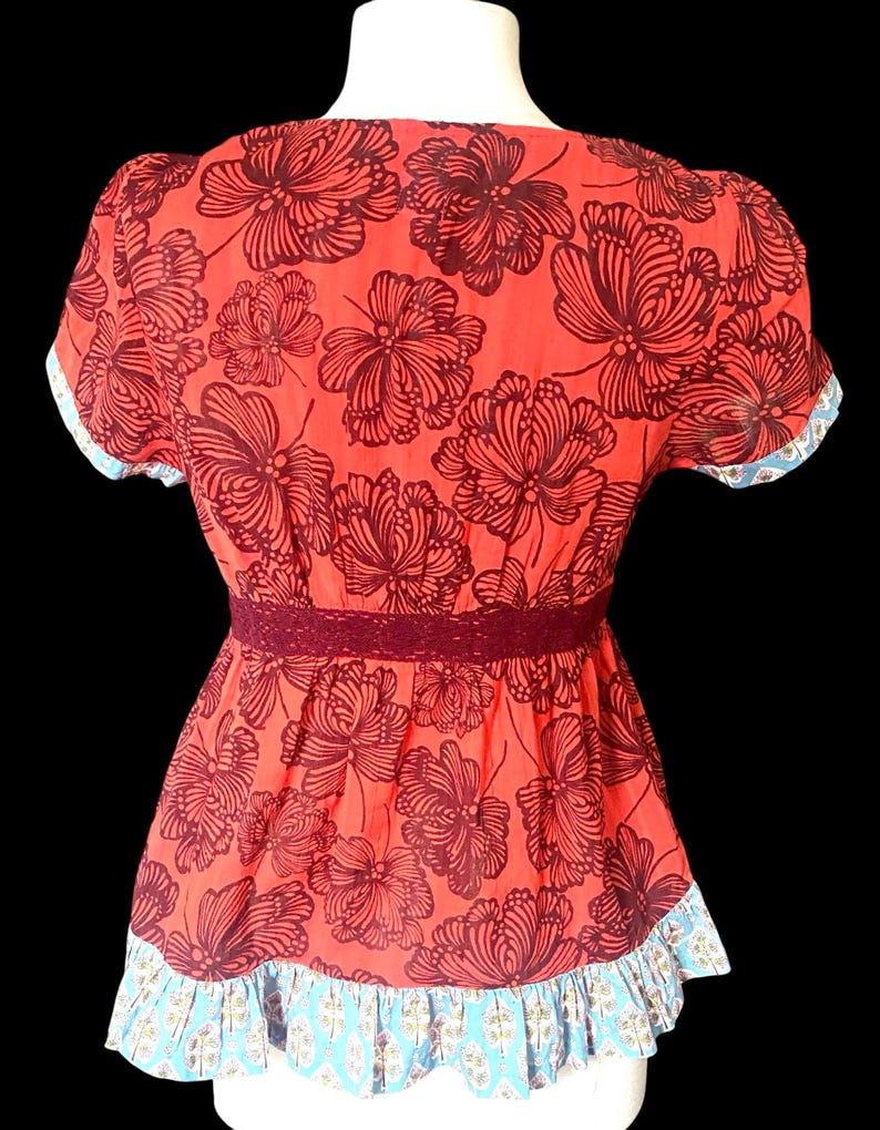 May include: A red floral top with a dark brown floral pattern. The top features short sleeves with a blue and white patterned trim, a burgundy lace waist detail, and a ruffled hem with a blue and white pattern. The top is displayed on a white mannequin.