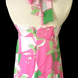 Op de afbeelding: Een halterjurk met een opvallende bloemenprint in roze, groen en wit. De jurk heeft een strik in de nek en een ritssluiting aan de achterkant. De jurk wordt op een mannequin gepresenteerd.