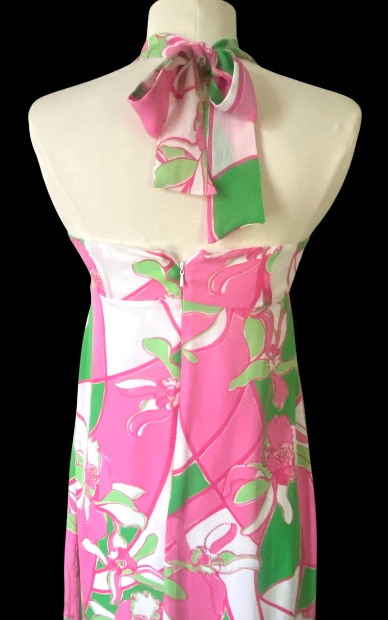 Op de afbeelding: Een halterjurk met een opvallende bloemenprint in roze, groen en wit. De jurk heeft een strik in de nek en een ritssluiting aan de achterkant. De jurk wordt op een mannequin gepresenteerd.