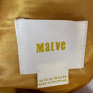 Op de afbeelding: Een wit rechthoekig kledinglabel met de merknaam "Maeve" in gele letters. Het label bevat maatdetails: US:12, UK:16, EU:44. Het label is geplaatst tegen een goudgele stoffen achtergrond.