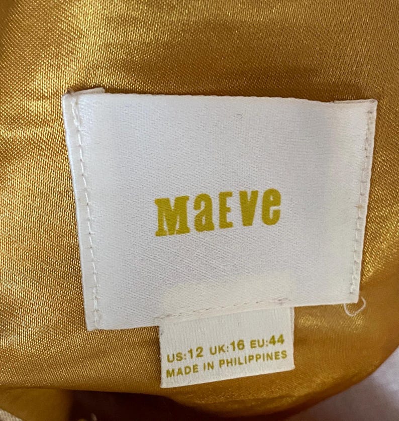Op de afbeelding: Een wit rechthoekig kledinglabel met de merknaam "Maeve" in gele letters. Het label bevat maatdetails: US:12, UK:16, EU:44. Het label is geplaatst tegen een goudgele stoffen achtergrond.