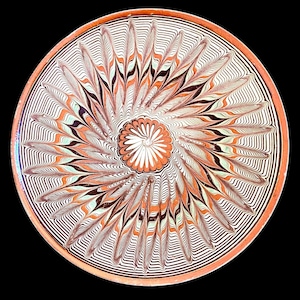 May include: A decorative ceramic plate with a detailed, symmetrical design. The plate features a central floral pattern in white and orange, surrounded by radiating leaf-like shapes in orange, black, and white. The rim is a warm orange color.
