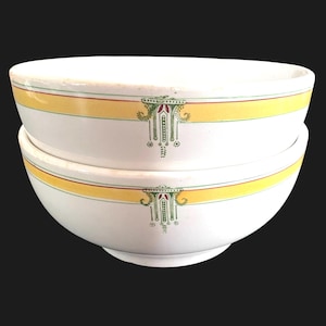May include: Two white ceramic bowls stacked on top of each other. Each bowl has a yellow band with a red stripe and green accents. A decorative green and red design is centered on each bowl.