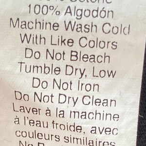 May include: A white fabric tag with black text detailing fabric composition and care instructions. The tag lists 100% cotton in multiple languages, including English, French, German, Spanish, and Italian. Washing instructions are also provided.