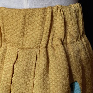 Op de afbeelding: Close-up van een mosterdgele rok met een textuur, honingraatachtig patroon. De rok is in de taille geplooid en heeft een geplooid ontwerp. Een klein gedeelte van lichtblauwe stof is zichtbaar.