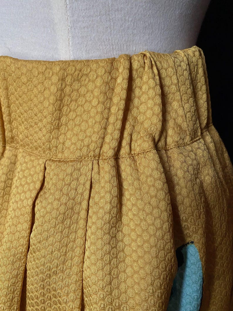 Op de afbeelding: Close-up van een mosterdgele rok met een textuur, honingraatachtig patroon. De rok is in de taille geplooid en heeft een geplooid ontwerp. Een klein gedeelte van lichtblauwe stof is zichtbaar.