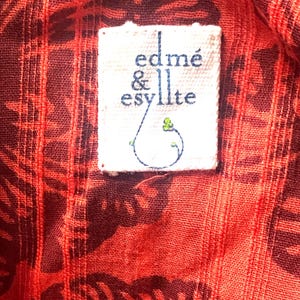 May include: Close-up of a red fabric with a dark pattern and a white label. The label reads "edm&eacute; & esyllte" in a handwritten style, with a small blue and green design. The fabric has a textured appearance.