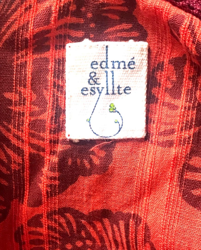 May include: Close-up of a red fabric with a dark pattern and a white label. The label reads "edm&eacute; & esyllte" in a handwritten style, with a small blue and green design. The fabric has a textured appearance.